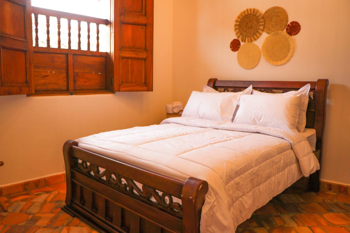 A neatly made bed with white bedding, a wooden headboard, and a window with wooden shutters.