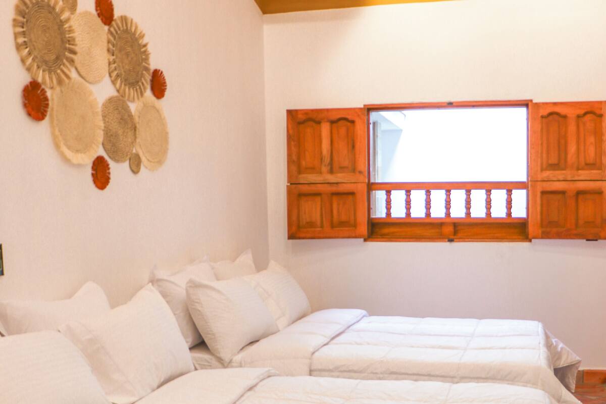 A bedroom with two beds, wooden headboards, a window with wooden shutters, and a hanging light fixture.
