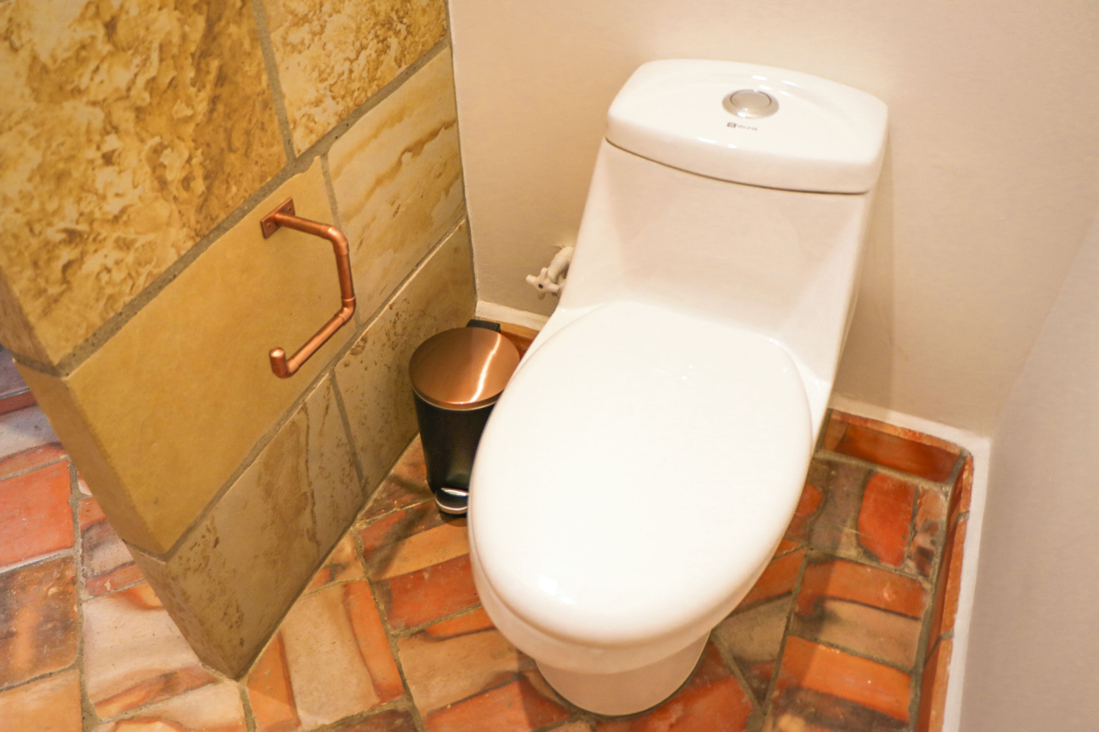 A toilet with a white seat and lid, a copper handrail, and a trash can with a black lid.