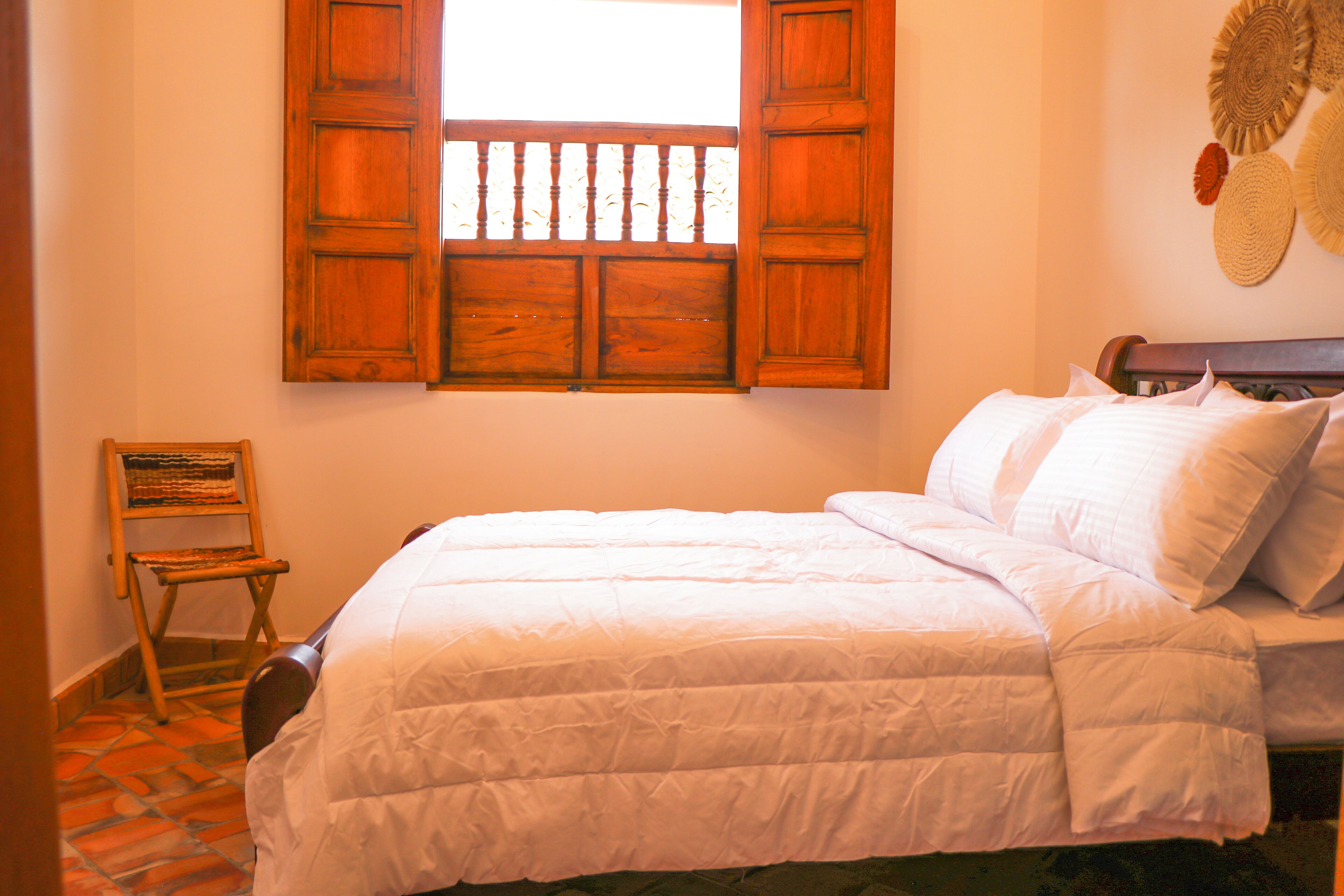 A bedroom with a bed, a wooden chair, a window with wooden shutters, and a wall with woven baskets.