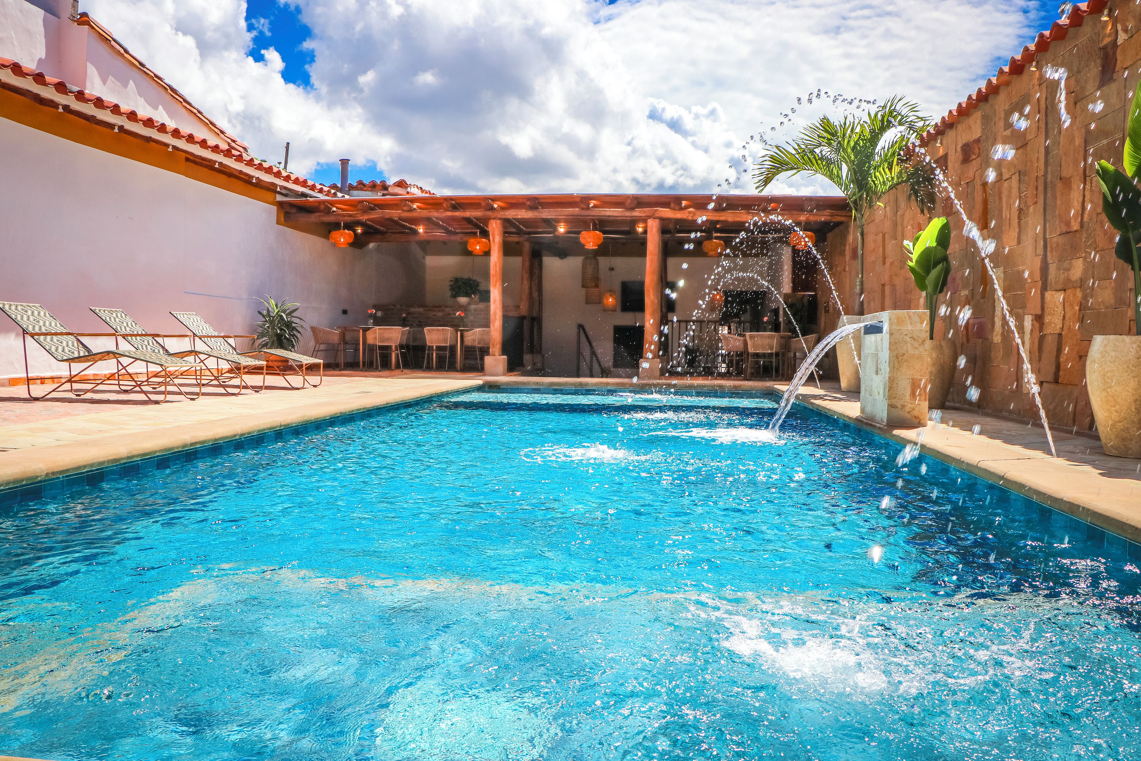Ein Schwimmbad mit Wasserspiel, Liegestühlen und einer überdachten Terrasse.
