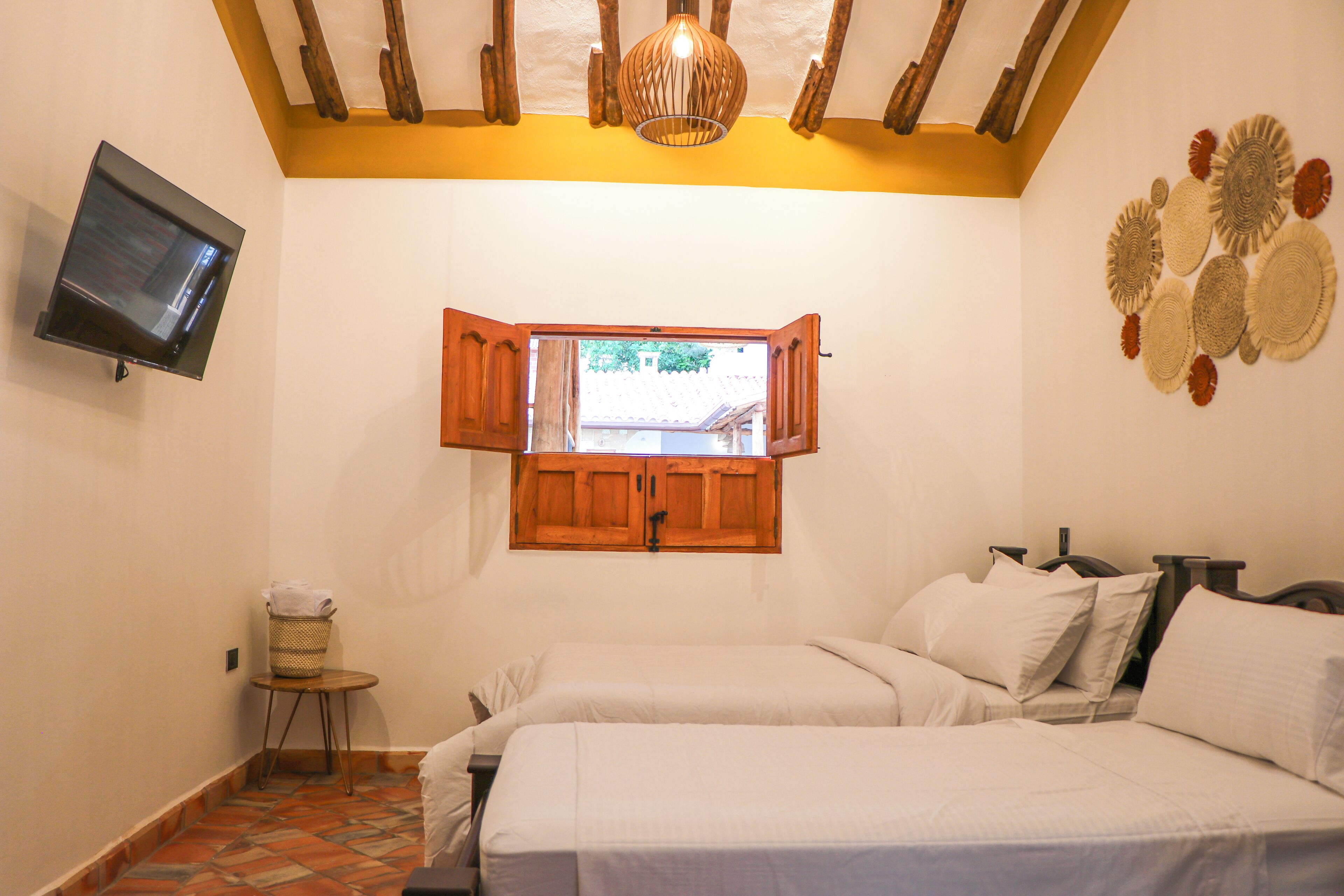 A bedroom with two beds, a television on the wall, a wooden ceiling, and a window with open shutters.