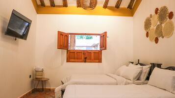 A bedroom with two beds, a television on the wall, a wooden ceiling, and a window with open shutters.