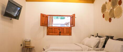 A bedroom with two beds, a television on the wall, a wooden ceiling, and a window with open shutters.