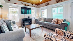 Ein gemütliches Wohnzimmer mit Holzbalkendecke, einem Flachbildfernseher, einem grauen Sofa, gemusterten Kissen und einem Couchtisch.