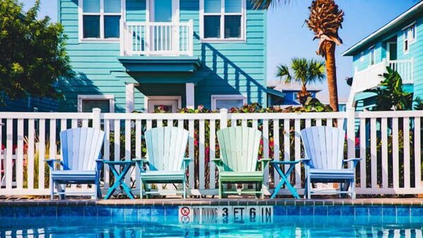 Ein Poolbereich mit weißem Zaun, ein blaues Haus mit Balkon und mehrere Adirondack-Stühle.