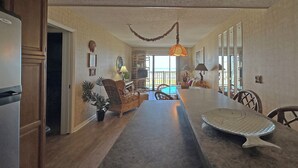 A spacious living area with a long dining table, wooden furniture, and a view to the outside. - Mariners Watch 101 - Oceanfront Complex in Garden City Beach (Garden City)