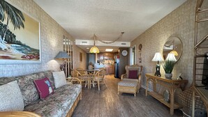A spacious living room with a sofa, dining table, and chairs, a kitchen in the background, and a large painting on the wall. - Mariners Watch 101 - Oceanfront Complex in Garden City Beach (Garden City)