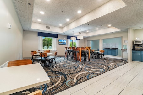 A hotel dining area with tables and chairs, a kitchen area with a microwave, and a large window with a view of trees.. The Windstar Hotel - Emerald Isle / Swansboro