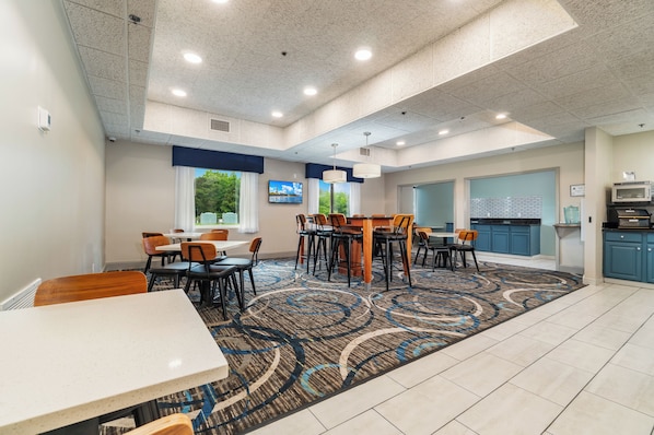 A hotel dining area with tables and chairs, a kitchen area with a microwave, and a large window with a view of trees.