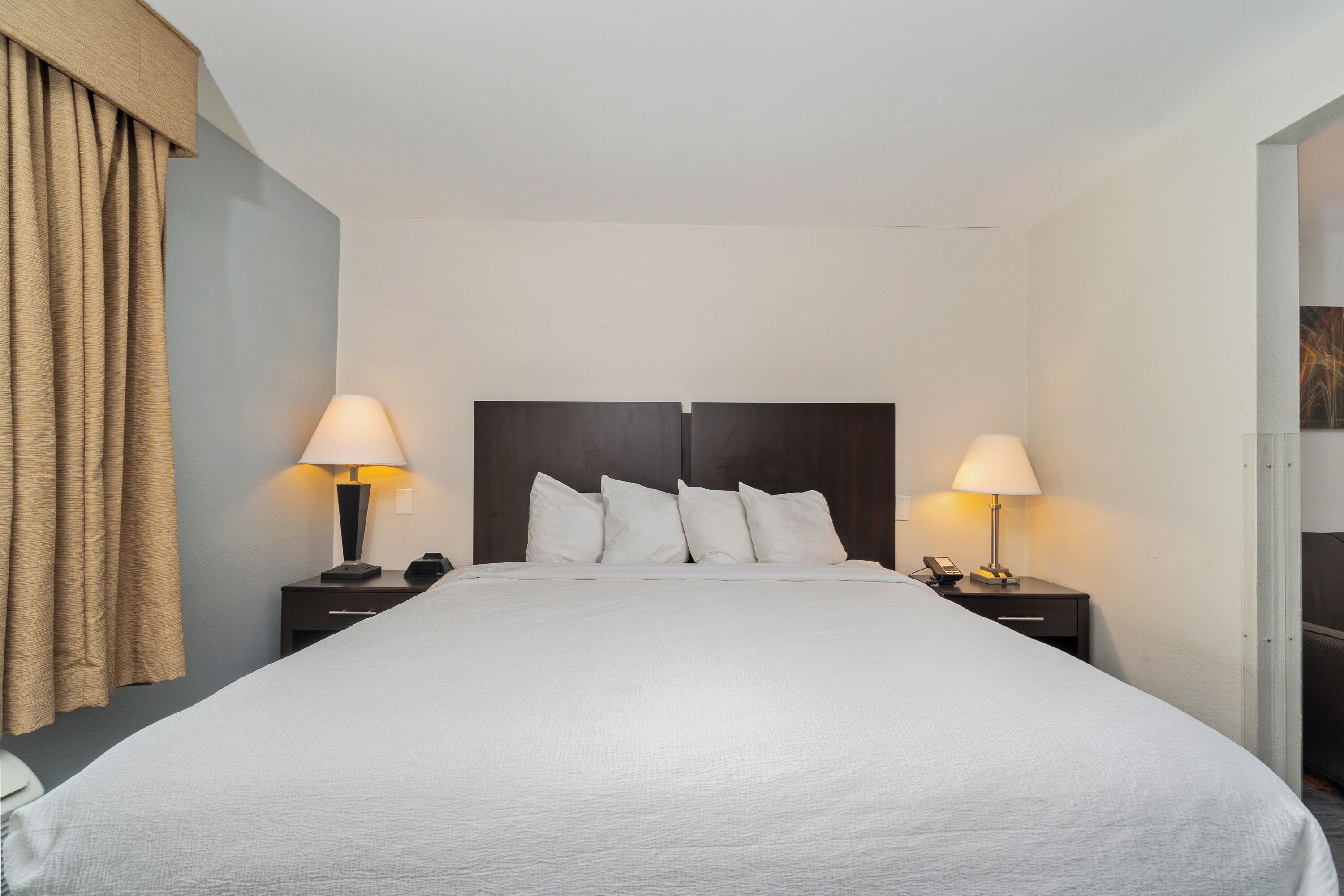 Une chambre d’hôtel avec un grand lit, deux tables de chevet avec des lampes, une tête de lit et un rideau sur le côté gauche.