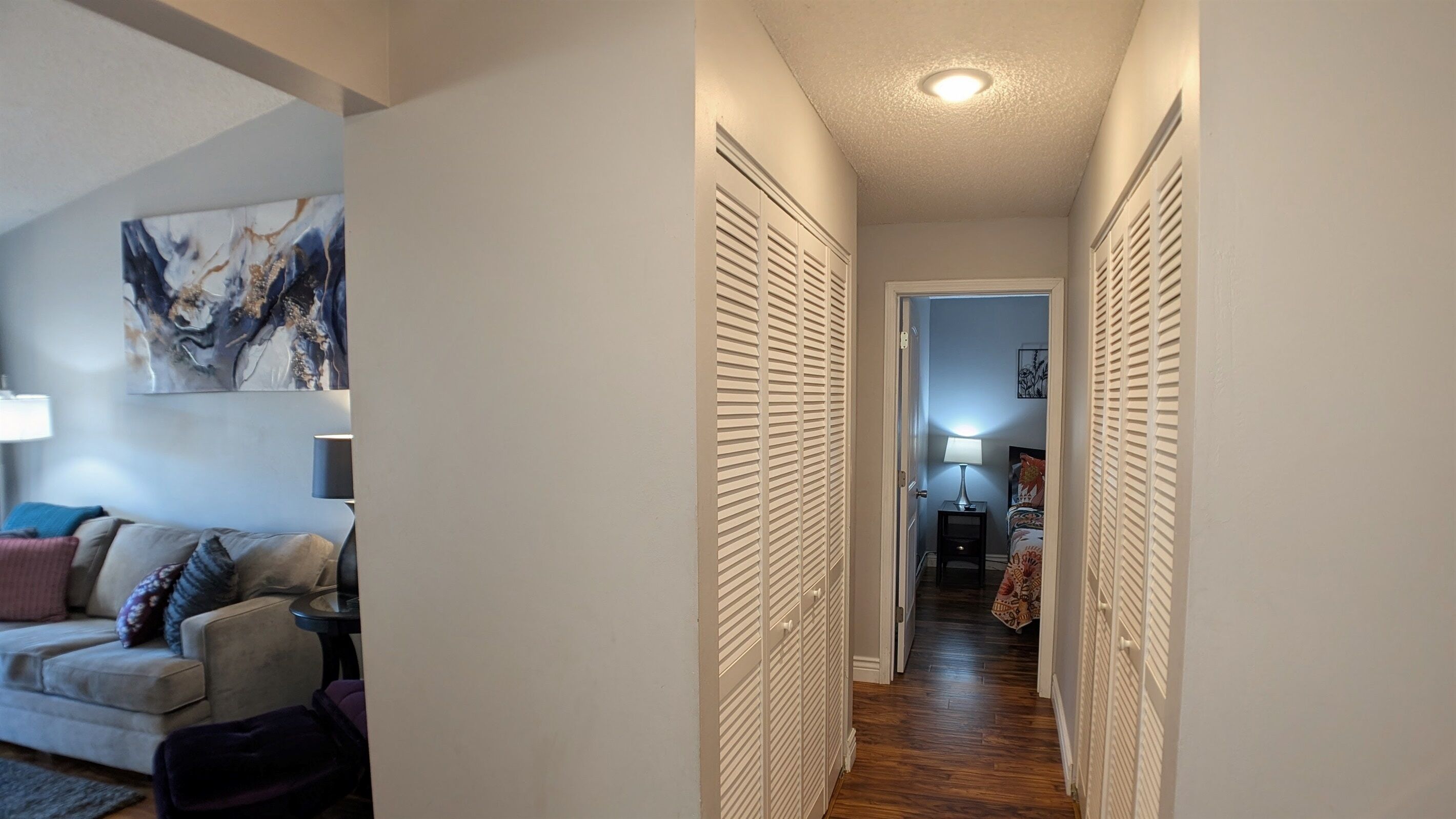 A narrow hallway with a ceiling light, leading to a bedroom with a bed and a nightstand.