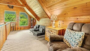 L’intérieur chaleureux d’un chalet en bois comprend une chambre à coucher située en mezzanine, un canapé, une table de chevet en bois et une vue sur la verdure environnante.