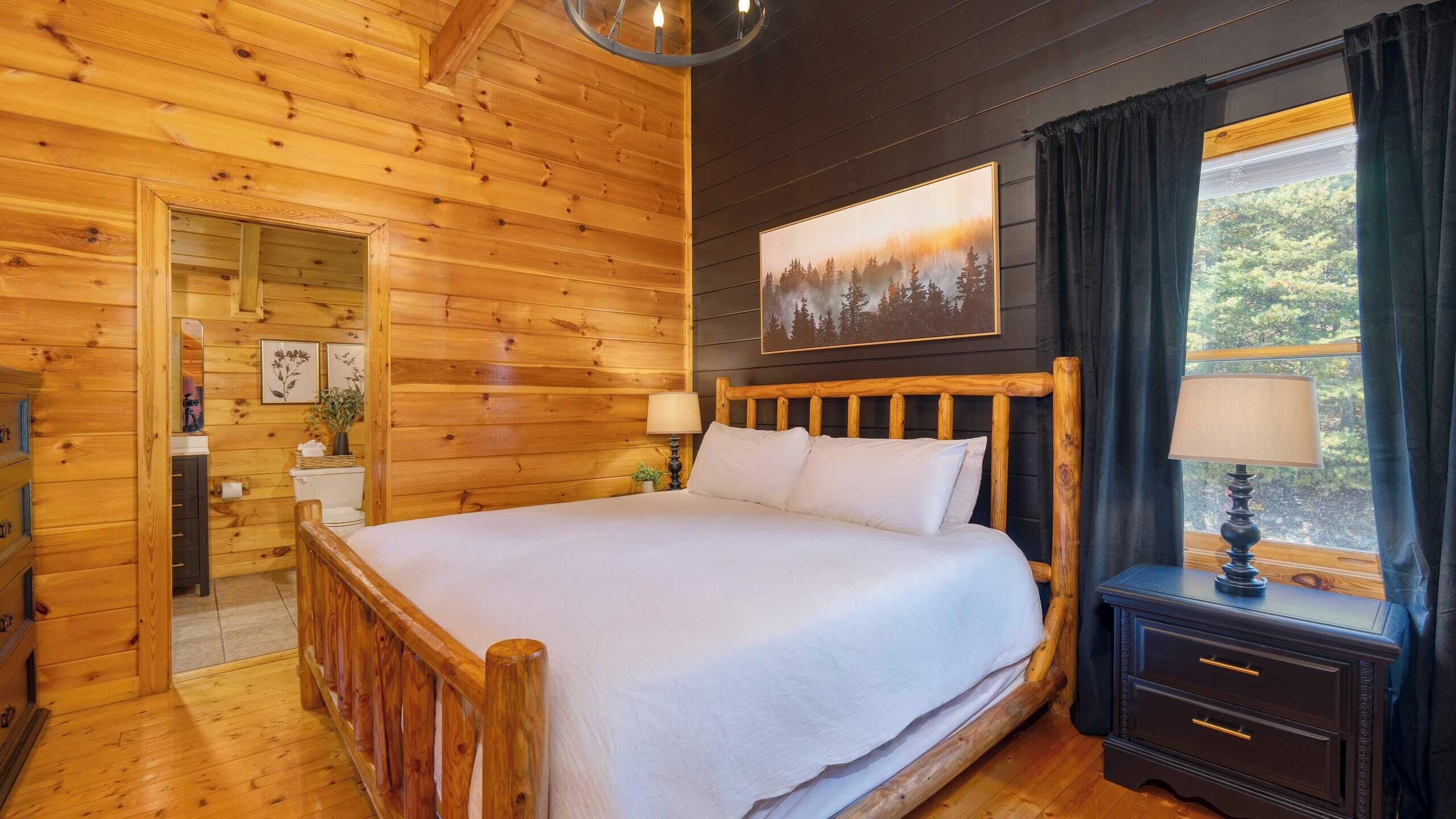 Een slaapkamer in een houten hutje met een bed, nachtkastjes, een raam met gordijnen en een schilderij aan de muur.