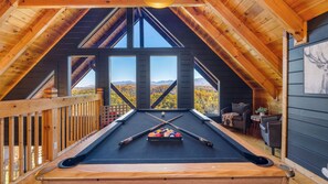 Une table de billard dans une pièce aux poutres apparentes, dotée de grandes fenêtres donnant sur un paysage montagneux.