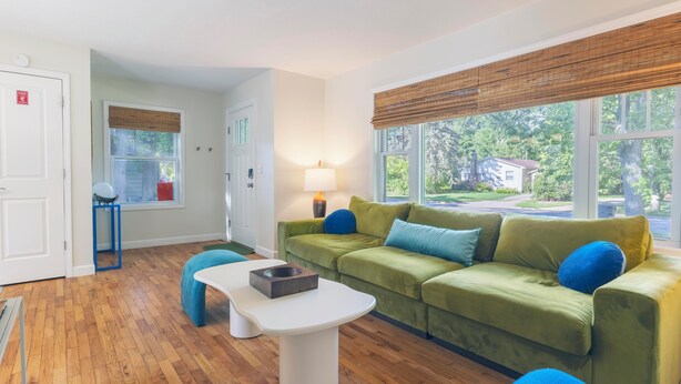 A modern living room with a green sofa, blue cushions, a white coffee table, and a lamp. There is a window with a view of a house and trees. - Retro Style Getaway- Game room- Hot tub (South Haven)