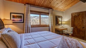 A bedroom with a large bed, a desk, a chair, a window with curtains, a framed picture, and a wooden door. - EagleVail Getaway | Ski, Golf & Hot Tub (Avon)