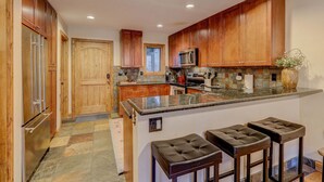 A modern kitchen with wooden cabinets, a granite countertop, and a built-in oven. There are three bar stools and a window above the sink. - EagleVail Getaway | Ski, Golf & Hot Tub (Avon)