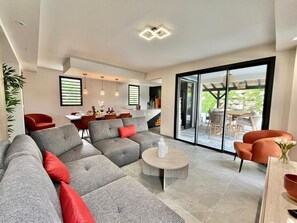 A modern living room with a grey sectional sofa, a round coffee table, and a dining area with a table and chairs. - Villa Aloha, Luxury in Martinique - Private Pool & 4 Suites in Trois-Îlets (Les Trois-Îlets)