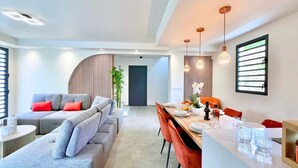 Een moderne woonkamer met een grijze modulaire bank, een eethoek met een houten tafel en rode stoelen, en een groot raam met zonwering.