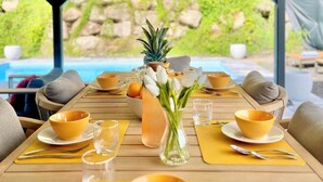 Een eethoek met een houten tafel gedekt voor vier personen, met gele kommen en glazen, een vaas met witte bloemen, en uitzicht op een zwembad en groen buiten.