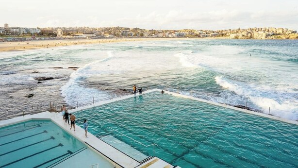 A coastal swimming pool with clear blue water, surrounded by a beach and a cityscape in the background. - KozyGuru | Waterloo | Designer Pad with City Views (Waterloo)