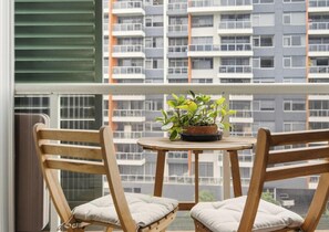En balkon med to træstole og et lille bord, en potteplante på bordet og udsigt til en høj beboelsesejendom.
