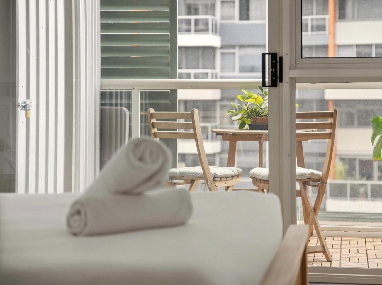 A hotel room with a bed, two wooden chairs, a small table with a plant, and a view of buildings through large windows.