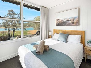 Un dormitorio con un gran ventanal que ofrece vistas a un lago y árboles. Hay una cama con cabecera, almohadas y una toalla enrollada sobre la cama.