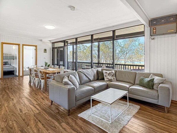 Una sala de estar moderna con un sofá seccional grande, una mesa de comedor con sillas y ventanas amplias que ofrecen vistas a árboles y un edificio.
