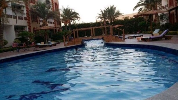 Une piscine entourée de chaises longues et de parasols, bordée de palmiers et d’un bâtiment doté de balcons.