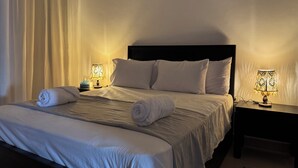 Un lit bien fait, avec du linge de lit blanc et deux serviettes enroulées, une table de chevet avec une lampe, et un rideau tiré partiellement sur le côté.