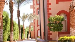 Un bâtiment à la façade rouge et beige, avec des palmiers et un petit arbre au premier plan.