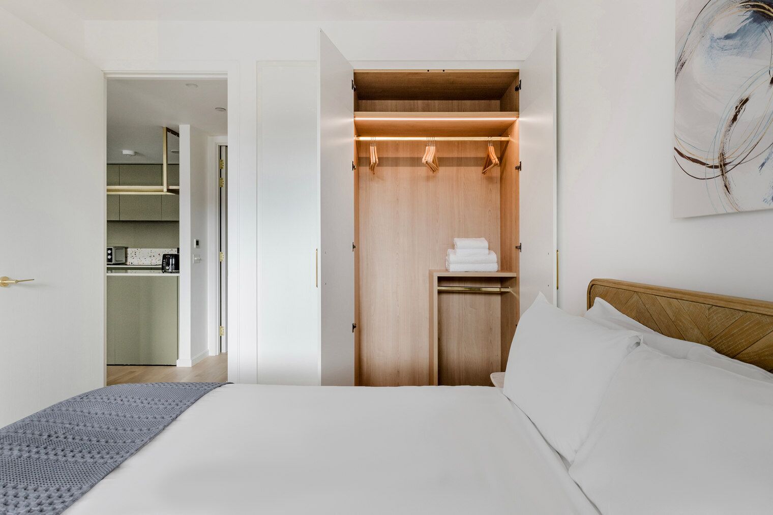 A modern bedroom with a wooden wardrobe, a bed with white linens, and a kitchen area visible through an open door.