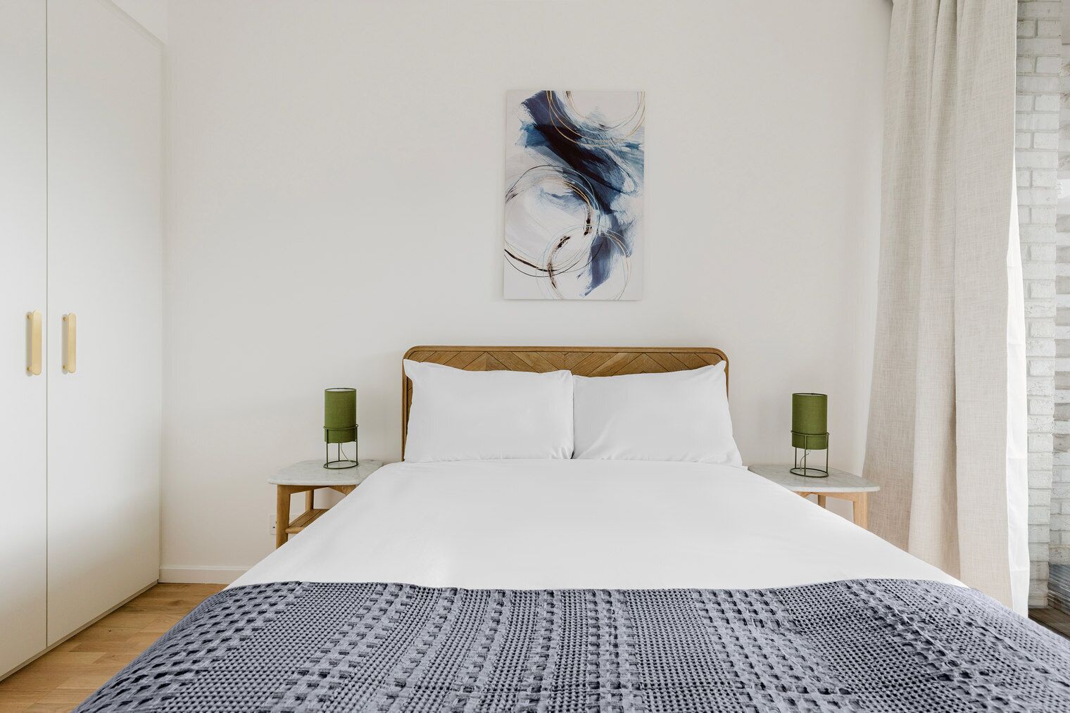 A neatly arranged bedroom with a bed, two bedside tables, a green lamp, and a large abstract painting on the wall.