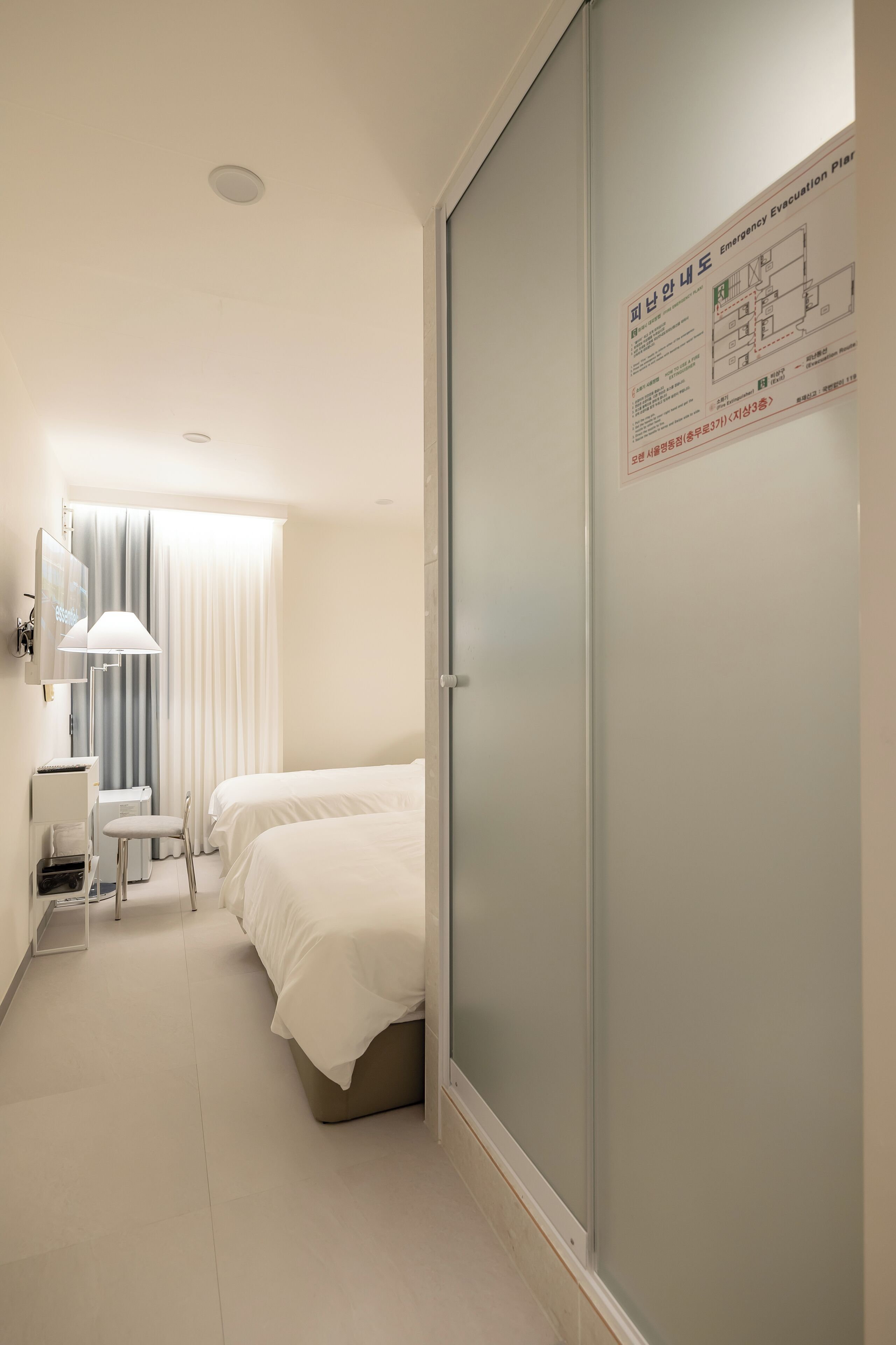 A hotel room with two beds, a desk, a chair, a lamp, and a wardrobe. There is a map on the wall and a window with curtains.