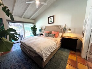 A bedroom with a bed, a nightstand, a lamp, a ceiling fan, a large plant, and a view of the outdoors. - Family Paradise! Ocean Views, Pool, Trampoline & Walk to Downtown (San Clemente)