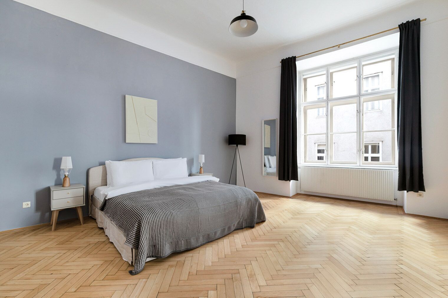 A bedroom with a bed, bedside tables, a floor lamp, a window with curtains, and a radiator.