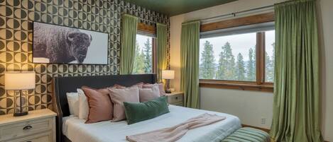 Un dormitorio con cama, mesitas de noche, lámpara, un cuadro en la pared y ventana con cortinas.