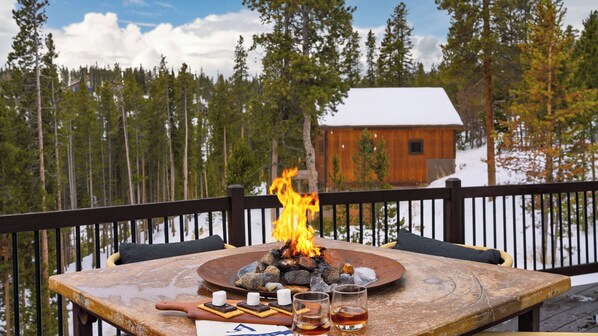 A wooden table with a fire pit, marshmallows, and glasses of a beverage, set against a snowy forest backdrop. - Skate Bowl, Climbing Wall, Arcade Breckenridge Adventure Chalet by Avantstay (Breckenridge)