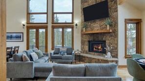 A spacious living room with a stone fireplace, large windows, and a flat-screen TV mounted on the wall. - Skate Bowl, Climbing Wall, Arcade Breckenridge Adventure Chalet by Avantstay (Breckenridge)