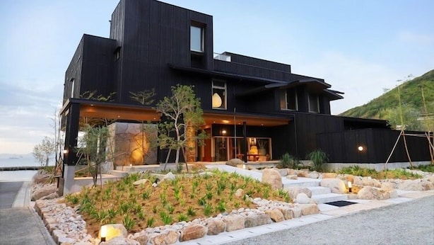 Una casa moderna de dos pisos con fachada oscura, cimientos de piedra y un jardín ajardinado con rocas y plantas.