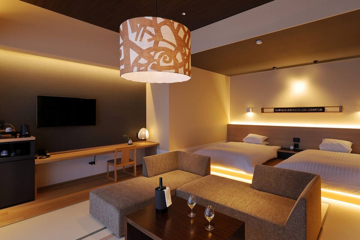 A hotel room with a bed, bedside tables, a TV, a desk, and a seating area with a lamp.