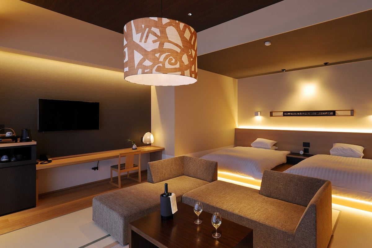 A hotel room with a bed, bedside tables, a TV, a desk, and a seating area with a lamp.