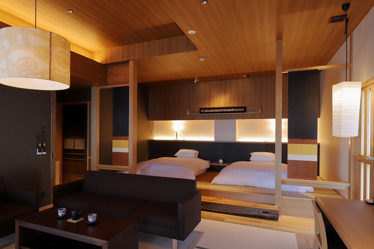 A modern hotel room with a wooden ceiling, two beds with headboards, a dark sofa, a small table with cups, and a large hanging light fixture.