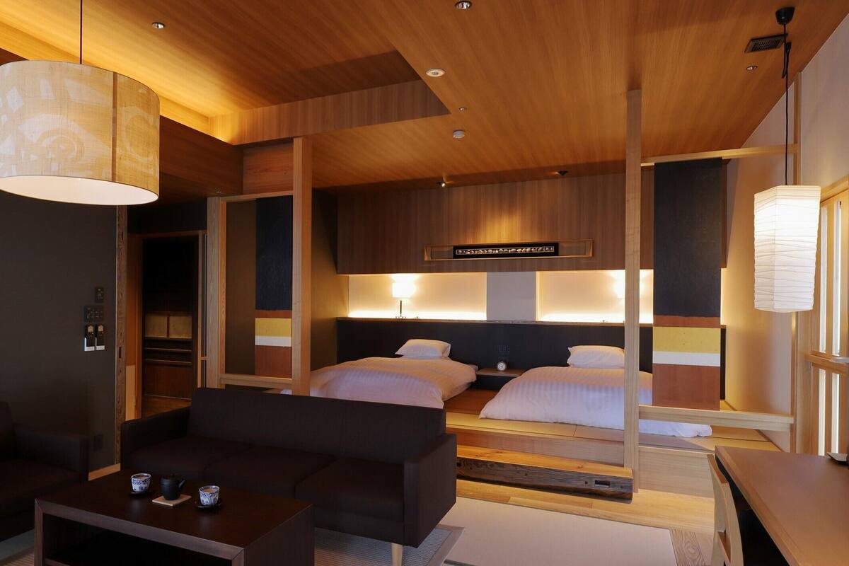 A modern hotel room with a wooden ceiling, two beds with headboards, a dark sofa, a small table with cups, and a large hanging light fixture.