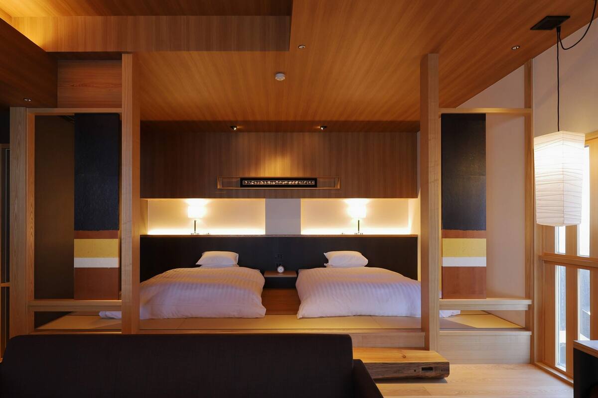 A hotel room with a wooden ceiling, two beds with white bedding, a nightstand with a clock, and a large window with a view.
