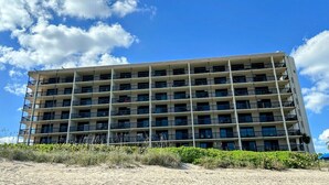 Un hotel a più piani con una serie di balconi, situato vicino a una spiaggia sabbiosa con vegetazione rada.
