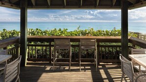 Un pontile in legno con due tavoli e quattro sedie, affacciato su una spiaggia con vista libera sull'oceano e una fascia di vegetazione.
