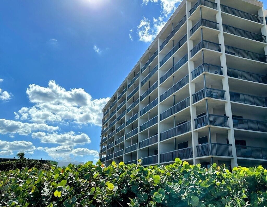 A multi-story hotel with balconies, a clear sky, and lush greenery in the foreground.